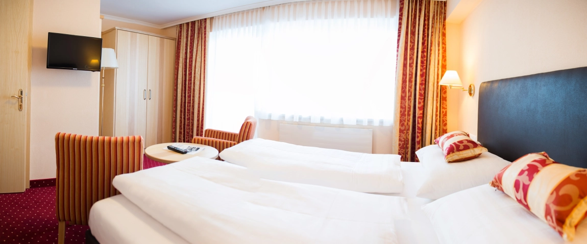 Helles Hotelzimmer mit zwei Einzelbetten, gemusterten Kissen, TV, Sitzbereich und großen Fenstern mit Vorhängen.